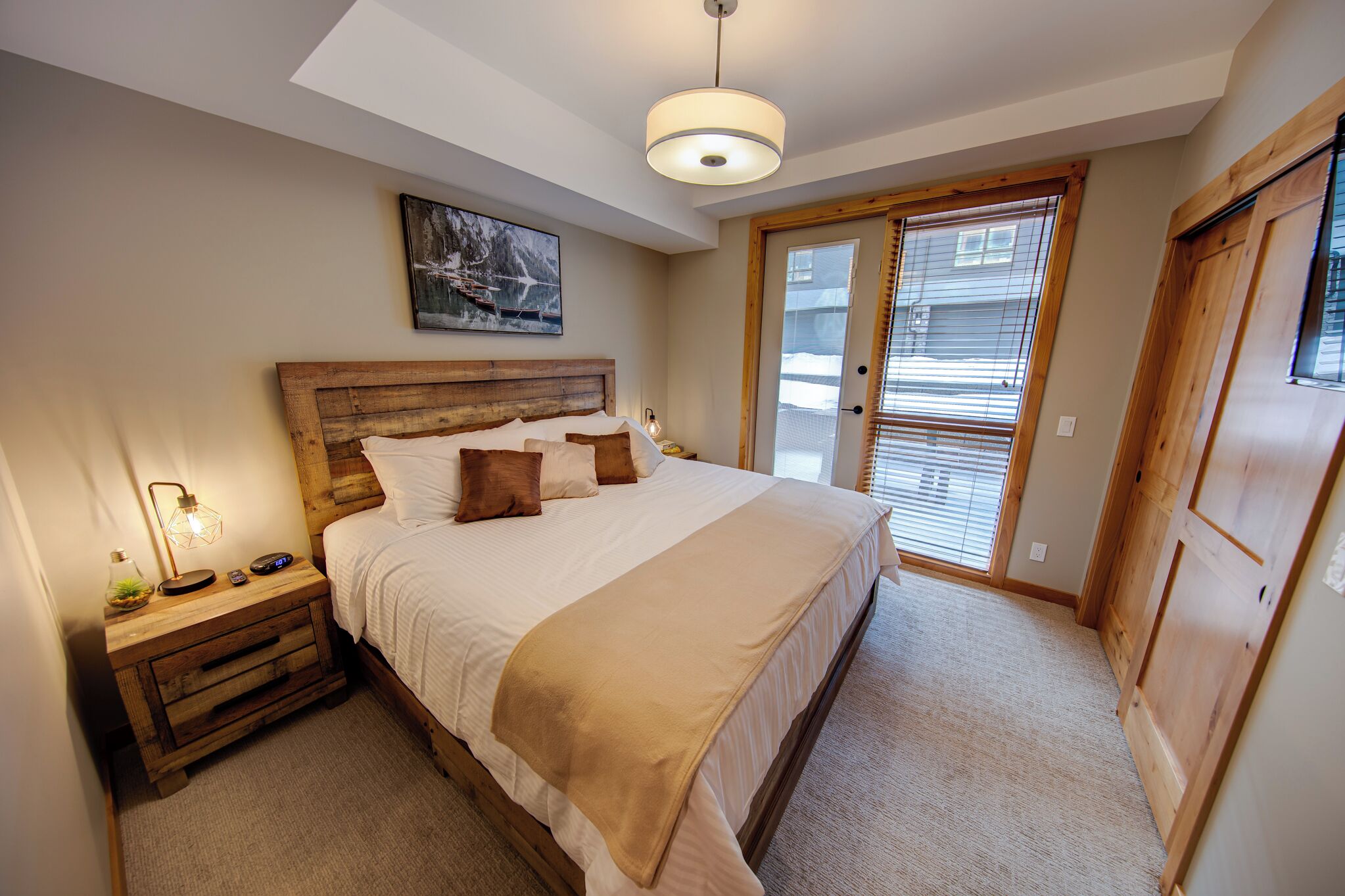 A bedroom with a wooden headboard, a large bed with white bedding, a bedside lamp, and a painting on the wall.