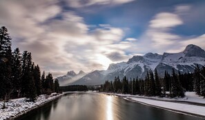 Ein Fluss mit schneebedeckten Ufern, umgeben von dichten Nadelbäumen und schneebedeckten Bergen unter einem teils bewölkten Himmel.