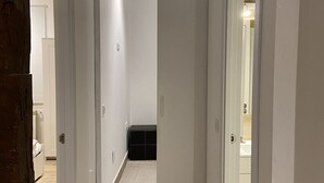 Un couloir étroit, doté d’un plancher en bois, mène à une chambre à coucher où se trouvent un lit et un petit pouf noir.