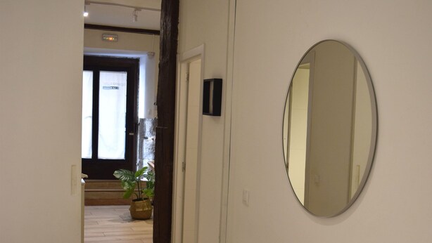 Une chambre d’hôtel avec un plancher en bois, un lit avec du linge de maison blanc, un miroir rond accroché au mur et une plante en pot dans un coin.