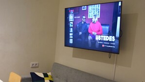 Un salon avec un canapé gris, une table blanche et un téléviseur fixé au mur, qui diffuse un programme d’information.