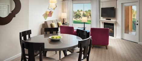 A hotel room with a round dining table, four chairs, a TV, a fireplace, and a view of a golf course.