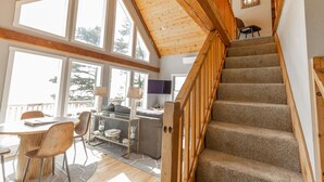 A spacious living area with a high ceiling, large windows, a staircase with wooden railings, and a cozy seating area with a sofa and chairs. - Honeymoon Cove at Crook Point (Brookings)
