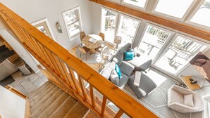 A spacious living area with a wooden staircase, a round table with chairs, a sofa with cushions, and large windows offering a view of the outdoors. - Honeymoon Cove at Crook Point (Brookings)