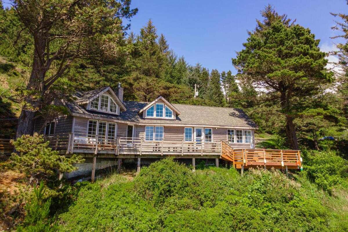 A wooden cabin with a deck, surrounded by dense forest.