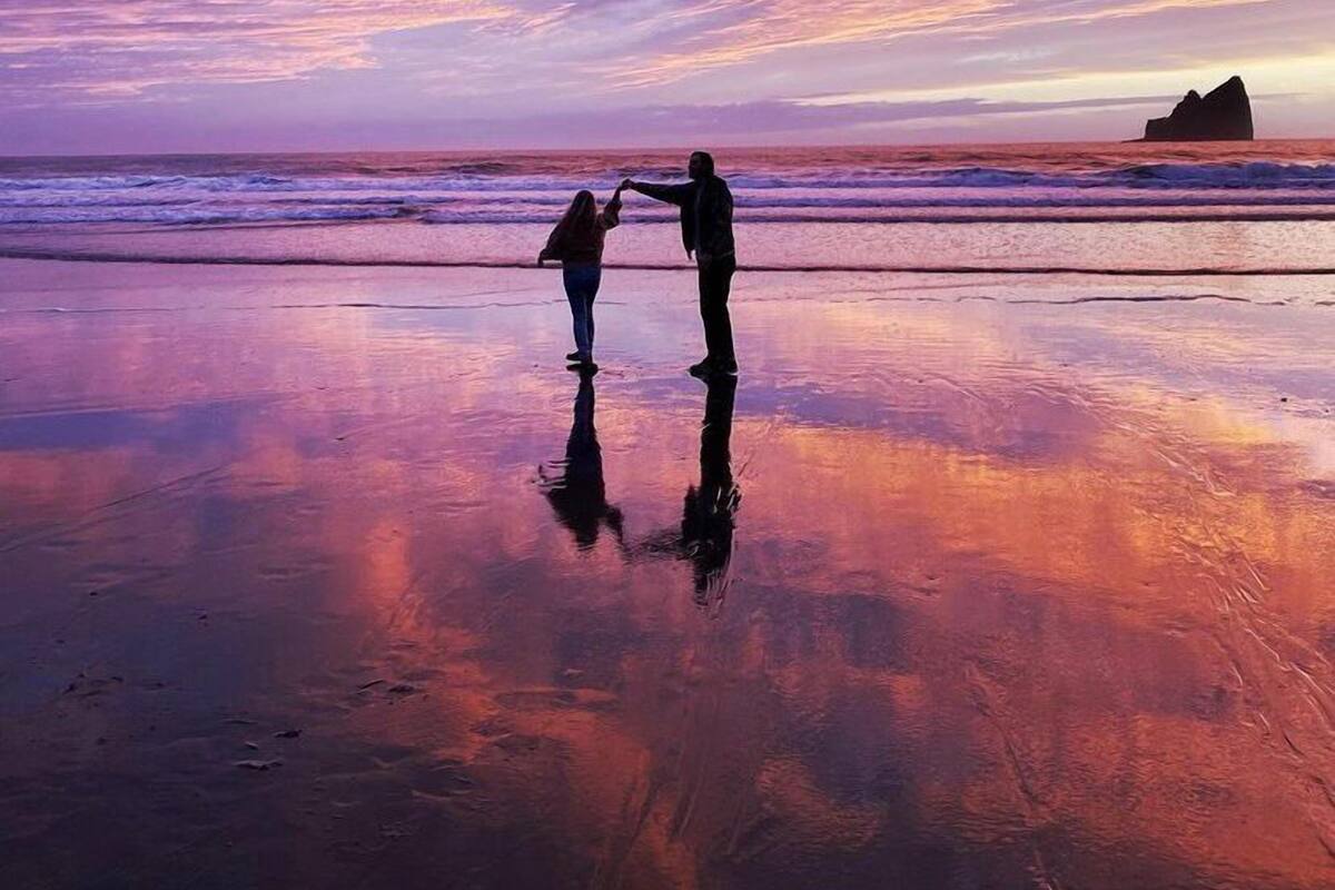 A beach scene with two people holding hands, a rocky outcrop in the distance, and a sunset sky.