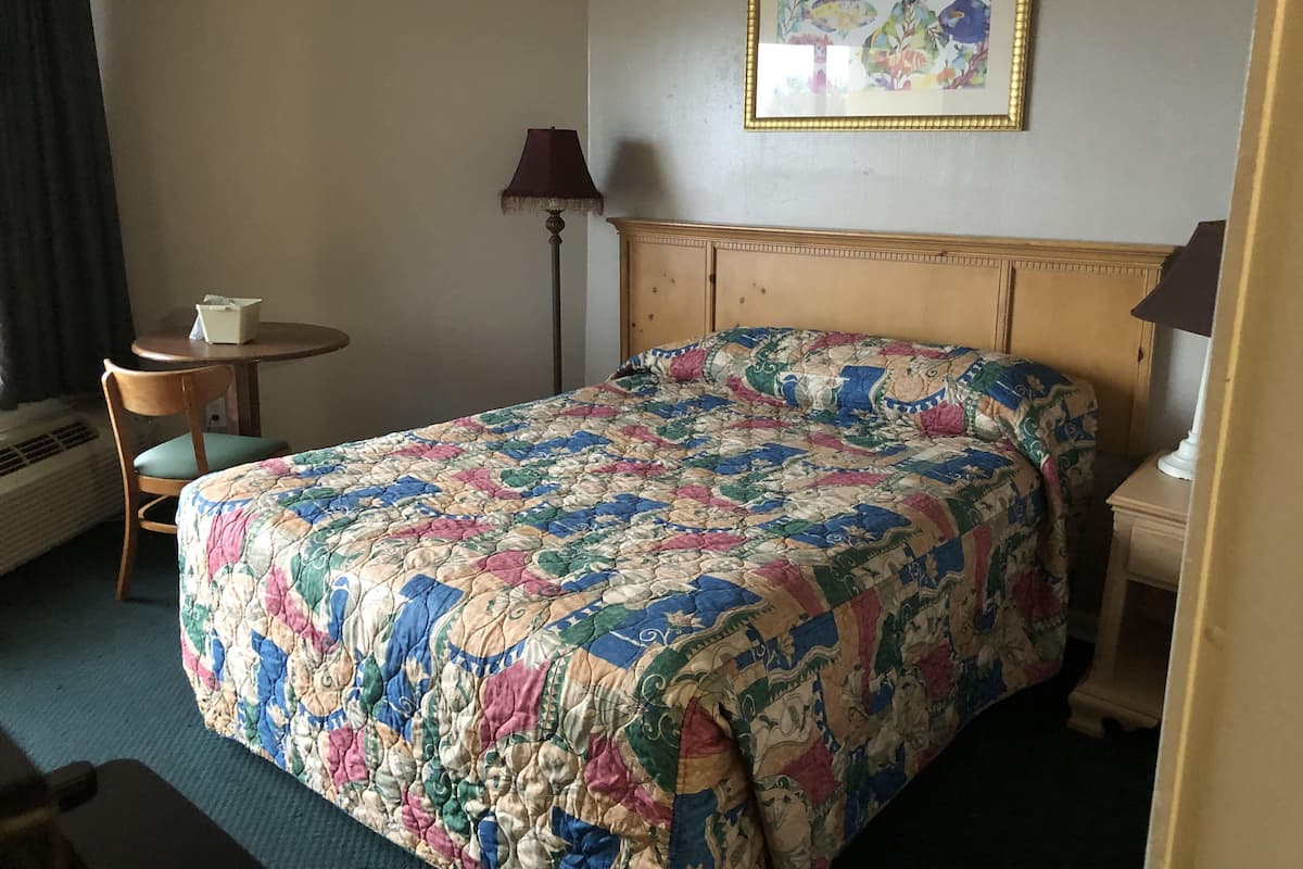 A hotel room with a bed, a small table, a chair, a lamp, and a painting on the wall.