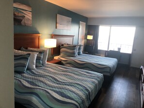 A hotel room with two beds, a nightstand, a lamp, a painting, and a window with curtains.