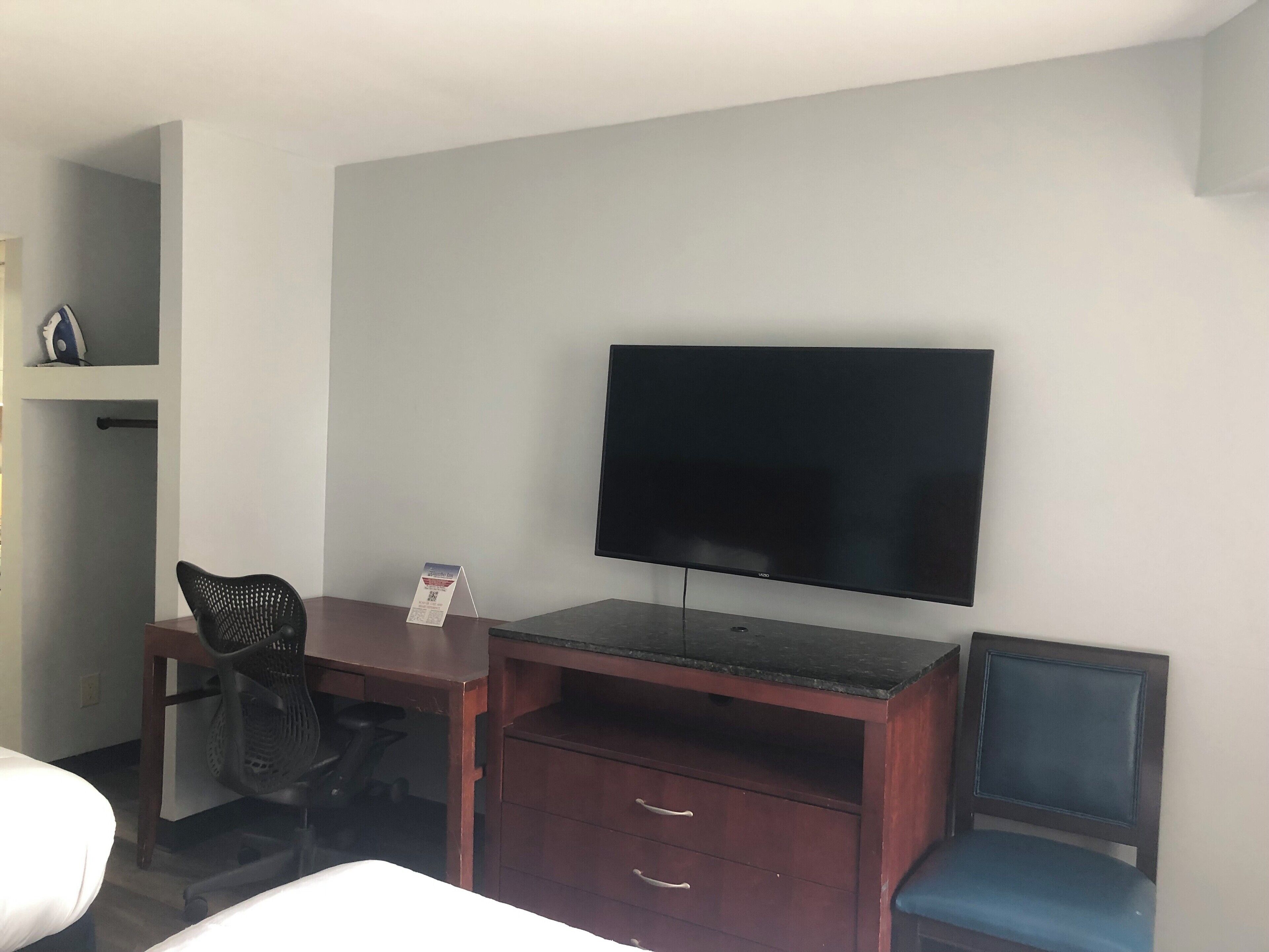 Camera d'albergo con televisione a schermo piatto montata a parete, una scrivania in legno con sedia, una piccola mensola con un oggetto decorativo e un letto singolo.