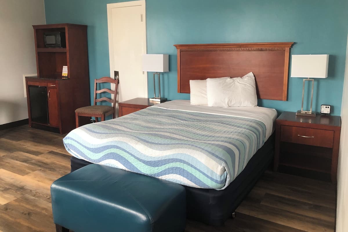 A hotel room with a bed, a desk with a microwave, a chair, a nightstand with a lamp, and a wooden cabinet.