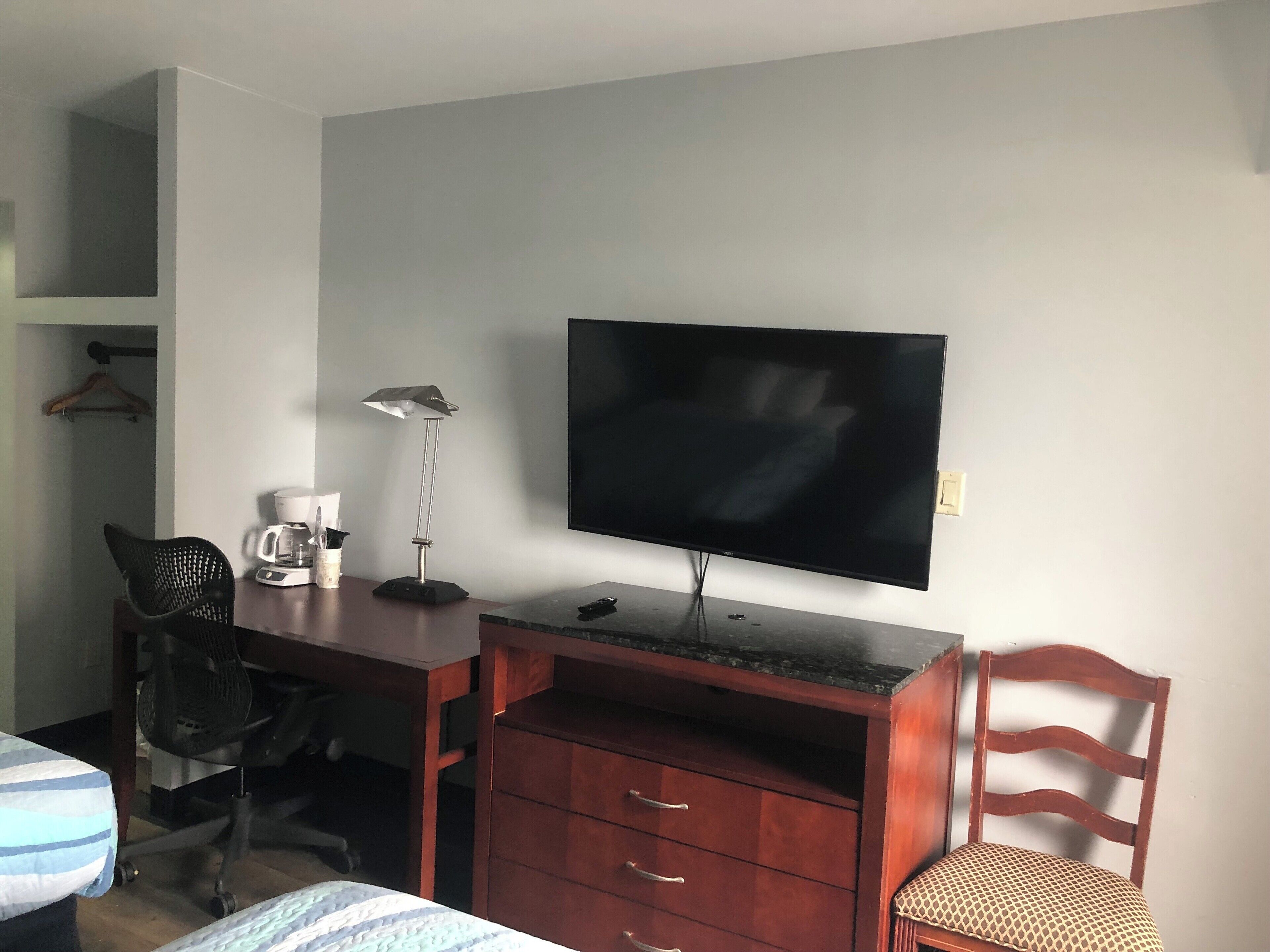 A hotel room with a wooden desk, a black office chair, a television mounted on the wall, a wooden dresser, and a bed with striped bedding.