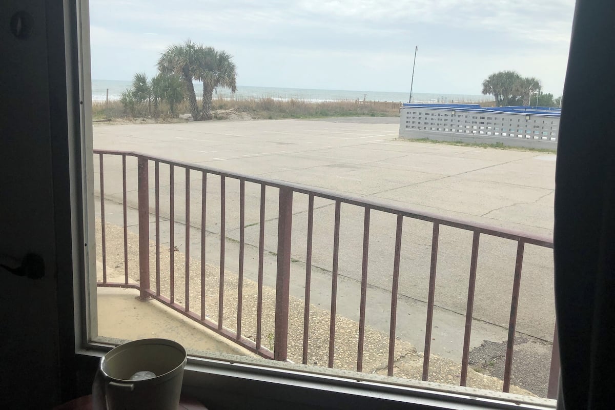 A view from a window with a metal railing, overlooking a parking area with a palm tree and a fence in the distance.