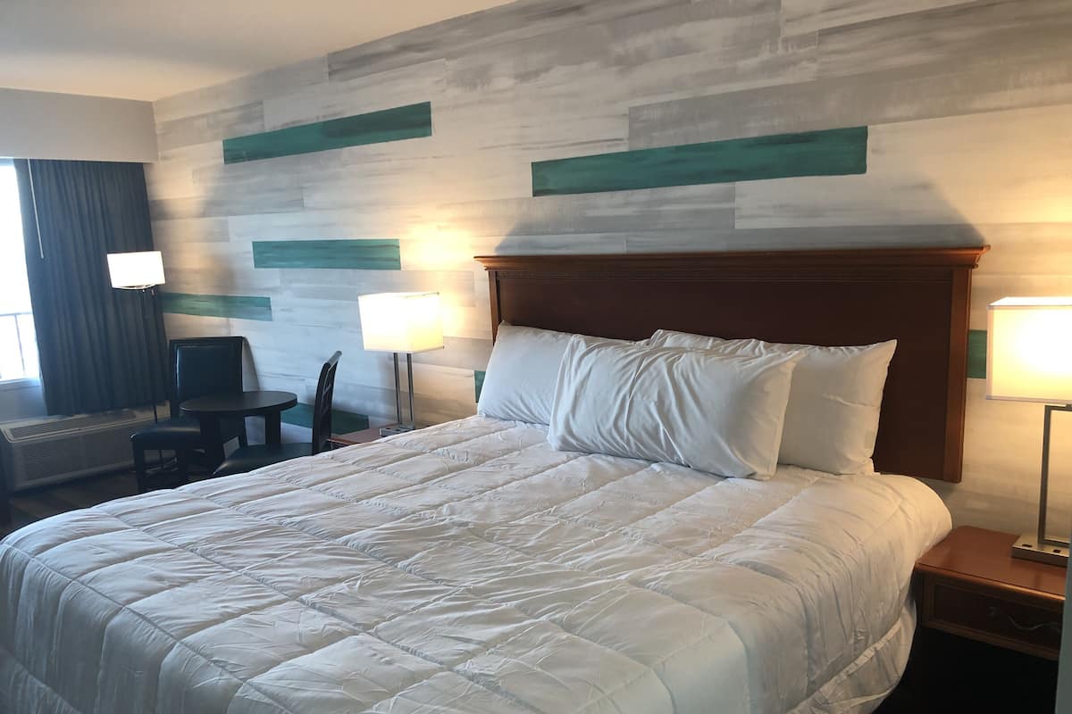 A hotel room with a large bed, a wooden headboard, two bedside lamps, a desk with a chair, and a window with curtains.