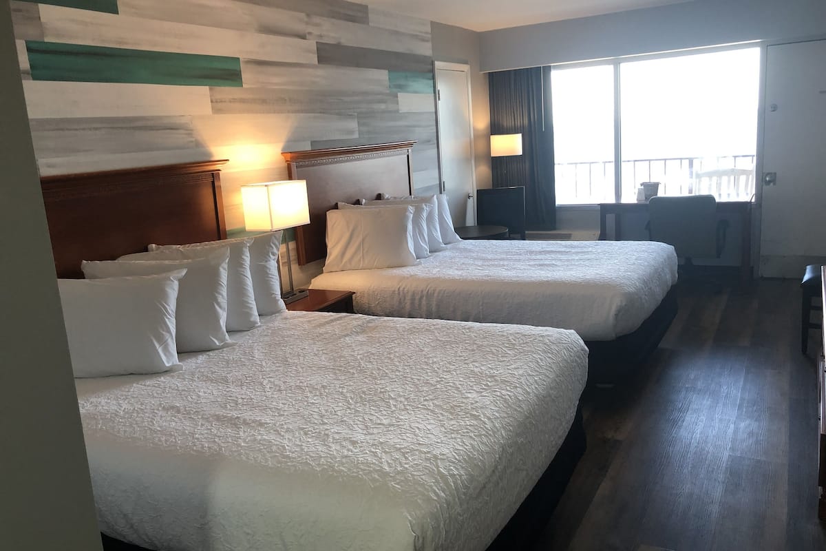 A hotel room with two beds, a desk, a chair, and a large window with curtains.