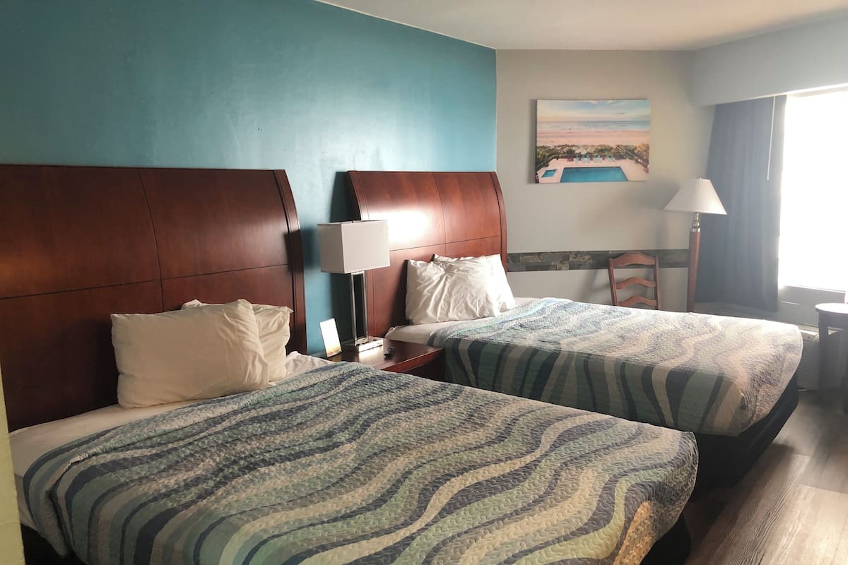 A hotel room with two beds, wooden headboards, a painting on the wall, a lamp, and a chair.