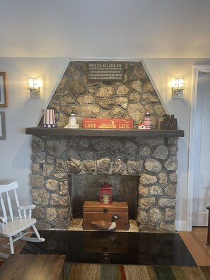 A stone fireplace with a sign above it, a wooden rocking chair, and a small table with a duck figurine.