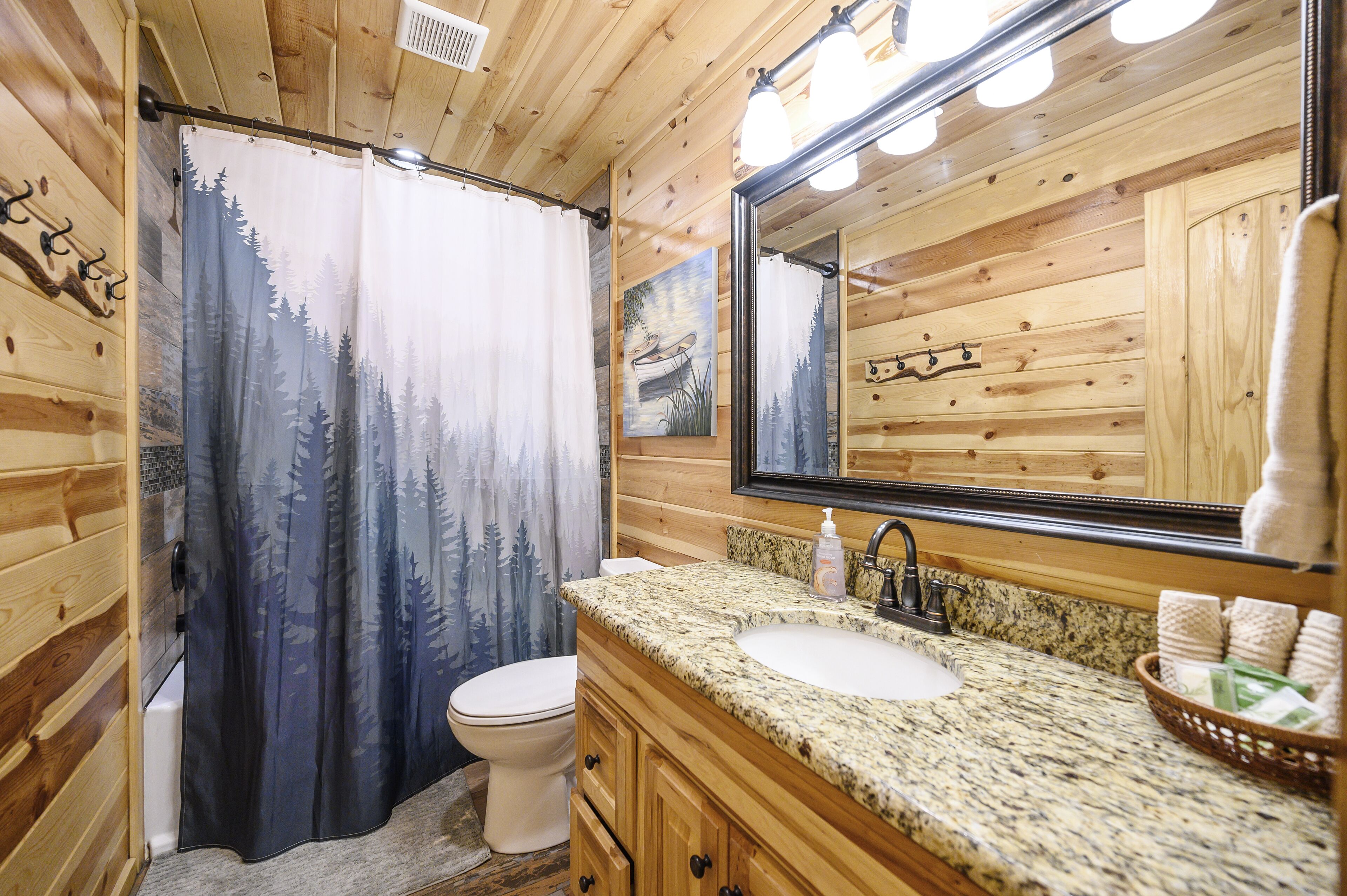 Une salle de bain avec un rideau de douche représentant une scène de forêt, des toilettes blanches, un lavabo sur un plan de travail en granit, un miroir et des meubles en bois.