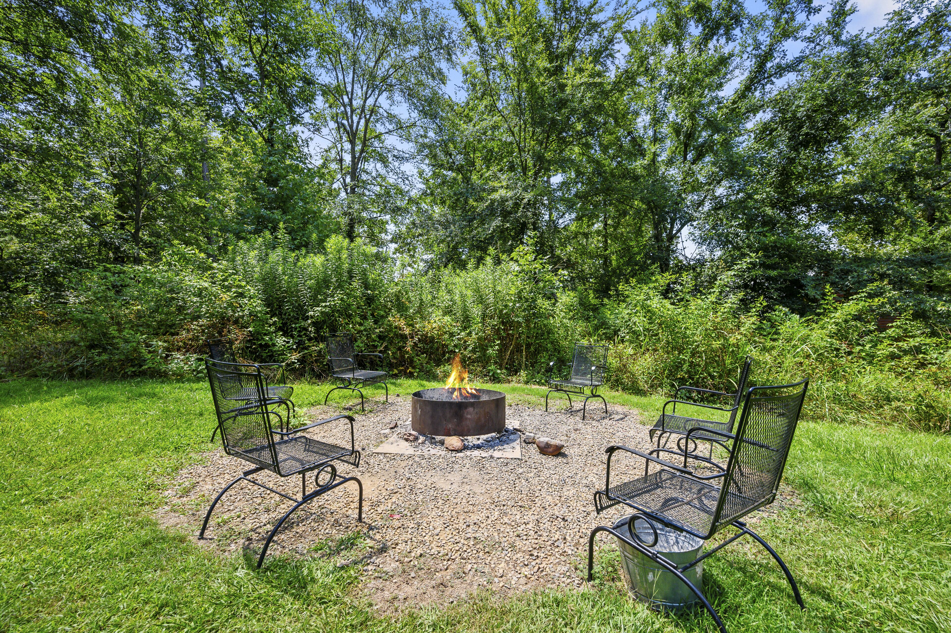 Een buitenruimte met zitplaatsen, een vuurkuil omringd door metalen stoelen, gelegen in een grasveld met bomen op de achtergrond.
