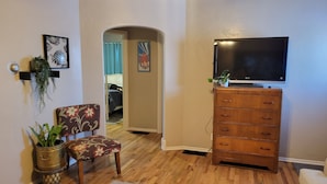 Una sala de estar con un aparador de madera, un televisor encima, una silla estampada, una planta pequeña y una alfombra.