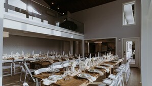 Une longue table à manger, dressée pour un événement formel, avec des chaises blanches, des tables en bois et des centres de table ornés de bougies.