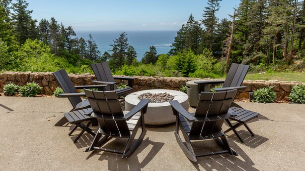 Un espace extérieur aménagé avec des chaises Adirondack et un foyer circulaire, offrant une vue sur un paysage boisé et l’océan.