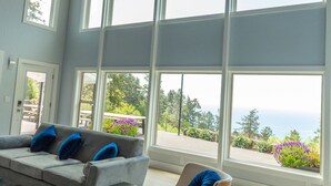Un salon spacieux avec un plafond haut, de grandes fenêtres, un canapé gris, un fauteuil beige et un tapis gris.