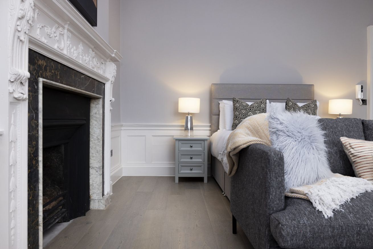 Un dormitorio con chimenea, una cama con cabecero gris, dos lámparas de noche y una mesita de noche con cajones.