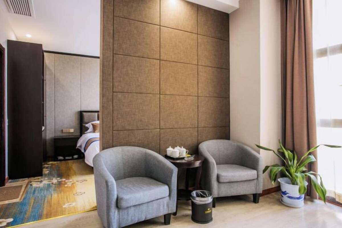 A modern hotel room with two grey armchairs, a small round table, a potted plant, and a large window with curtains.