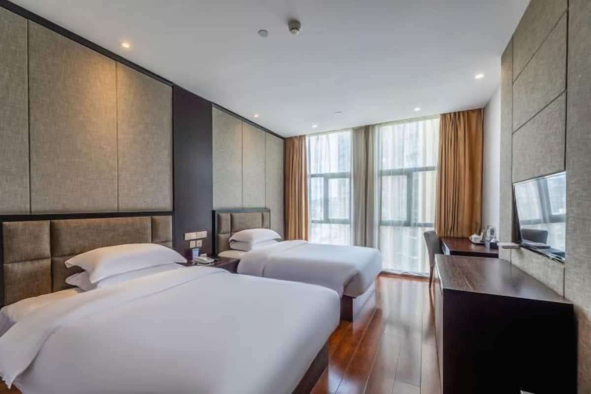 A hotel room with two beds, a desk, a chair, and large windows.