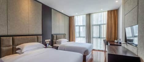 A hotel room with two beds, a desk, a chair, and large windows.