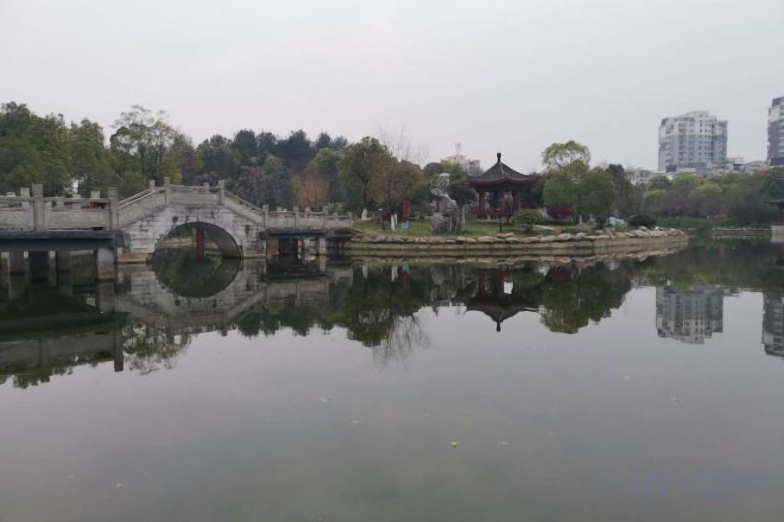 A tranquil park with a stone bridge, a pavilion, and a reflection in the water.