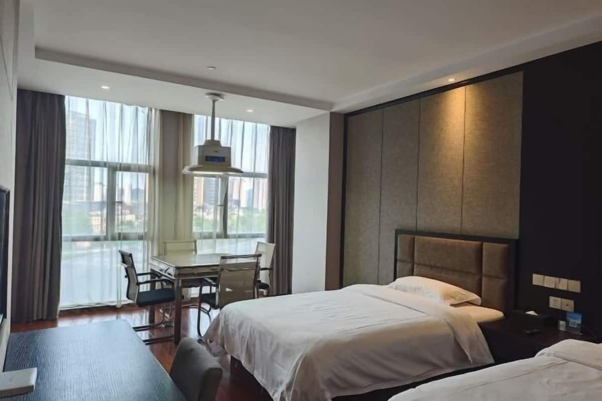 A hotel room with two beds, a desk with chairs, a large window with curtains, and a modern headboard.