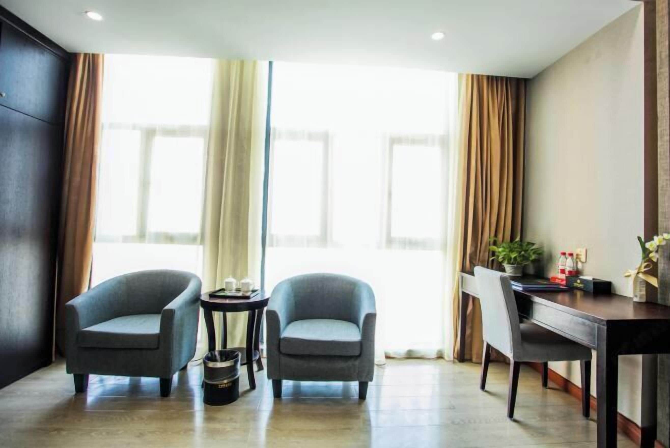 A hotel room with two armchairs, a small table, a desk, and a large window with curtains.