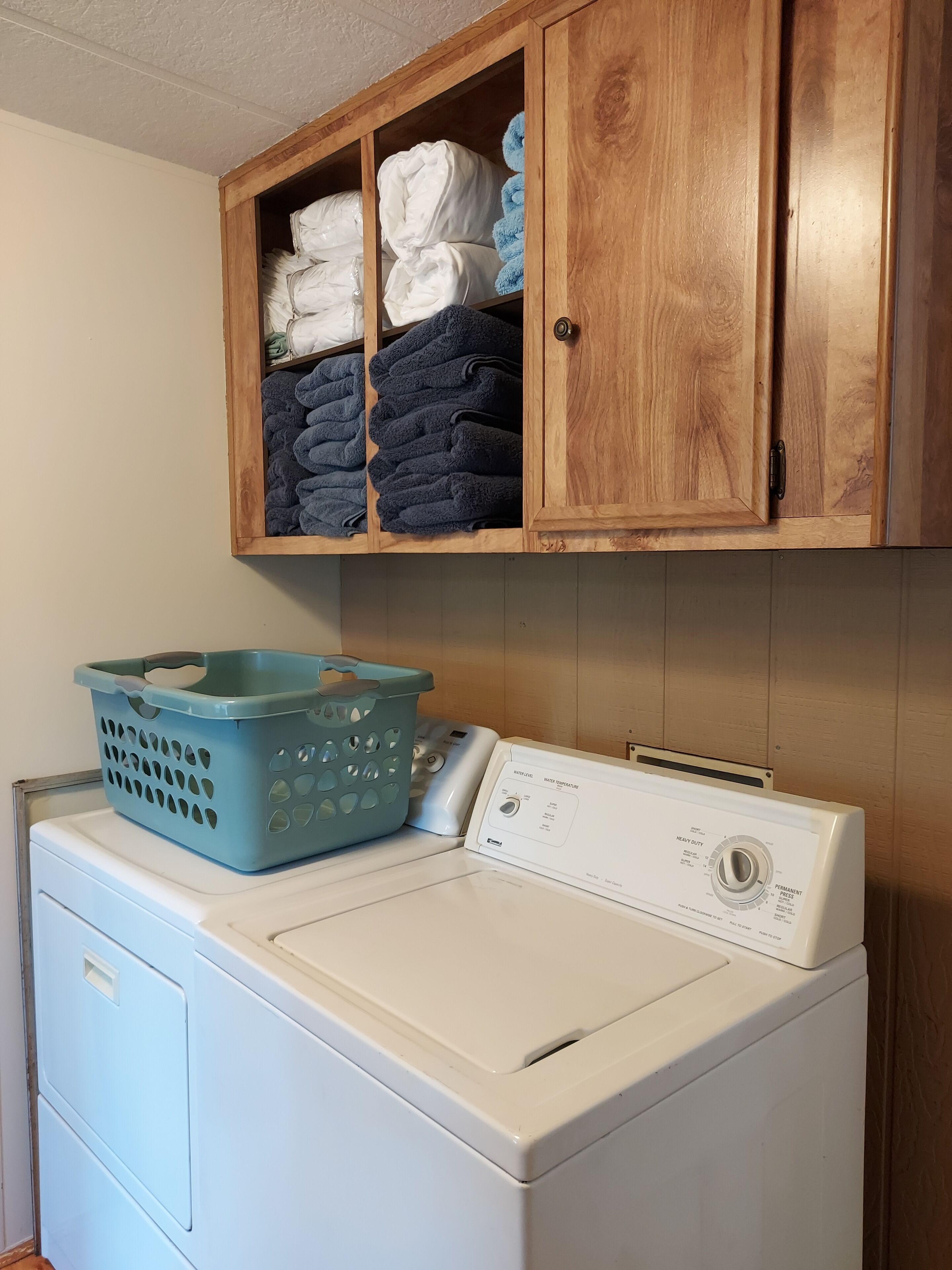 A front-loading washing machine with a blue laundry basket on top, a wooden cabinet above with folded towels and blankets, and a white wall.