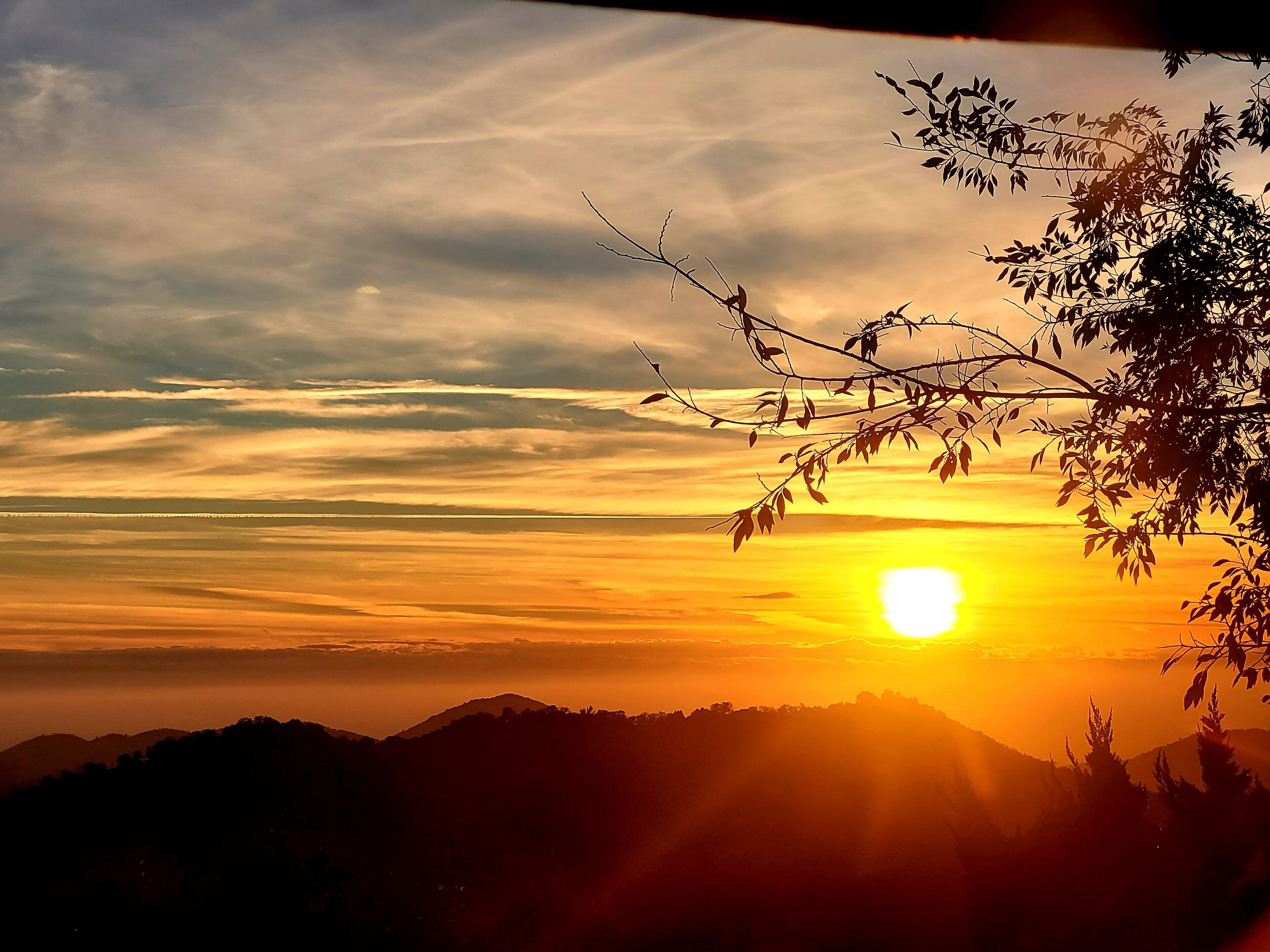 A sunset over a mountainous landscape with silhouettes of trees and hills.