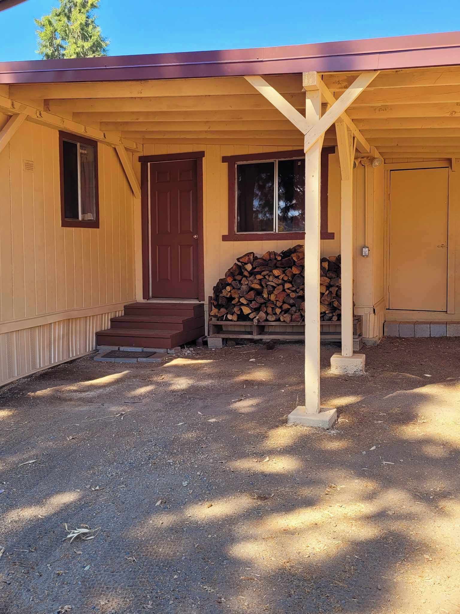 A single-story building with a brown door and windows, a covered porch, and a stack of firewood.