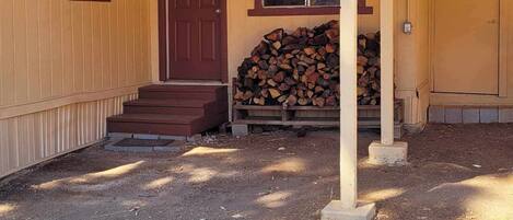 A single-story building with a brown door and windows, a covered porch, and a stack of firewood.
