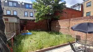 A backyard with a grassy area, a wooden fence, a tree, and a patio with a table and chairs. - Privet apartment with 2 bedrooms for relaxing escape in EastHam near CentralPark (London)