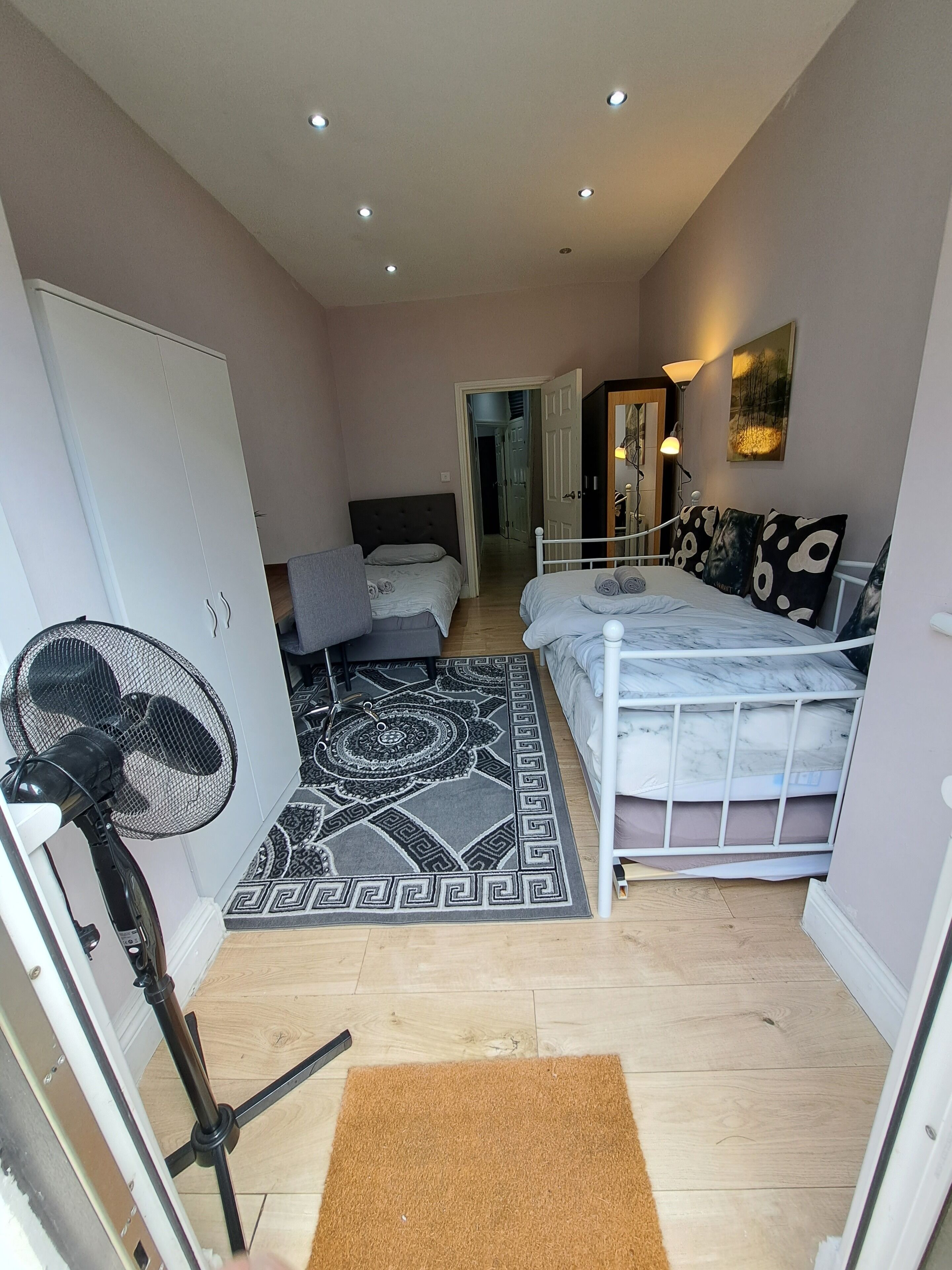 Ein gemütliches Schlafzimmer mit Bett, Standventilator, einem gemusterten Teppich und einer Tür zu einem anderen Raum.