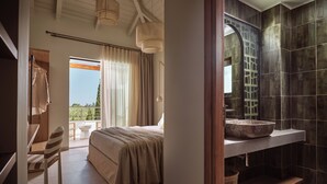 Une chambre avec un grand lit, une salle de bain avec des carreaux foncés et un grand lavabo, et un balcon avec des rideaux.