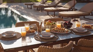 Un buffet de petit-déjeuner installé au bord de la piscine, avec une variété de fruits, de viennoiseries et de boissons disposés sur une table, entourée de chaises longues et offrant une vue sur la piscine.