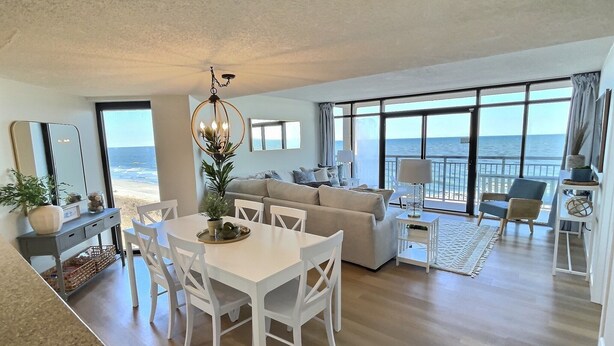 A spacious living area with a dining table, white chairs, a sofa, and a large window offering a view of the ocean. - Oceanfront Escape! Stylish 3 bedroom corner unit (North Myrtle Beach)