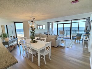 Amplia sala de estar con una mesa de comedor, sillas blancas, un sofá y un gran ventanal con vista al océano.