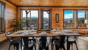 A wooden-paneled dining room with a large table set for six, large windows offering a view of the mountains, and a potted plant on the left. - Beautiful Mt. View Pool Cabin with Hot Tub & Arcade Games! Perfect for Families! (Sevierville)