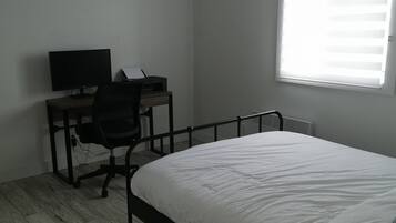 Une chambre à coucher avec un lit, un bureau avec un ordinateur, une chaise et une fenêtre avec des stores.