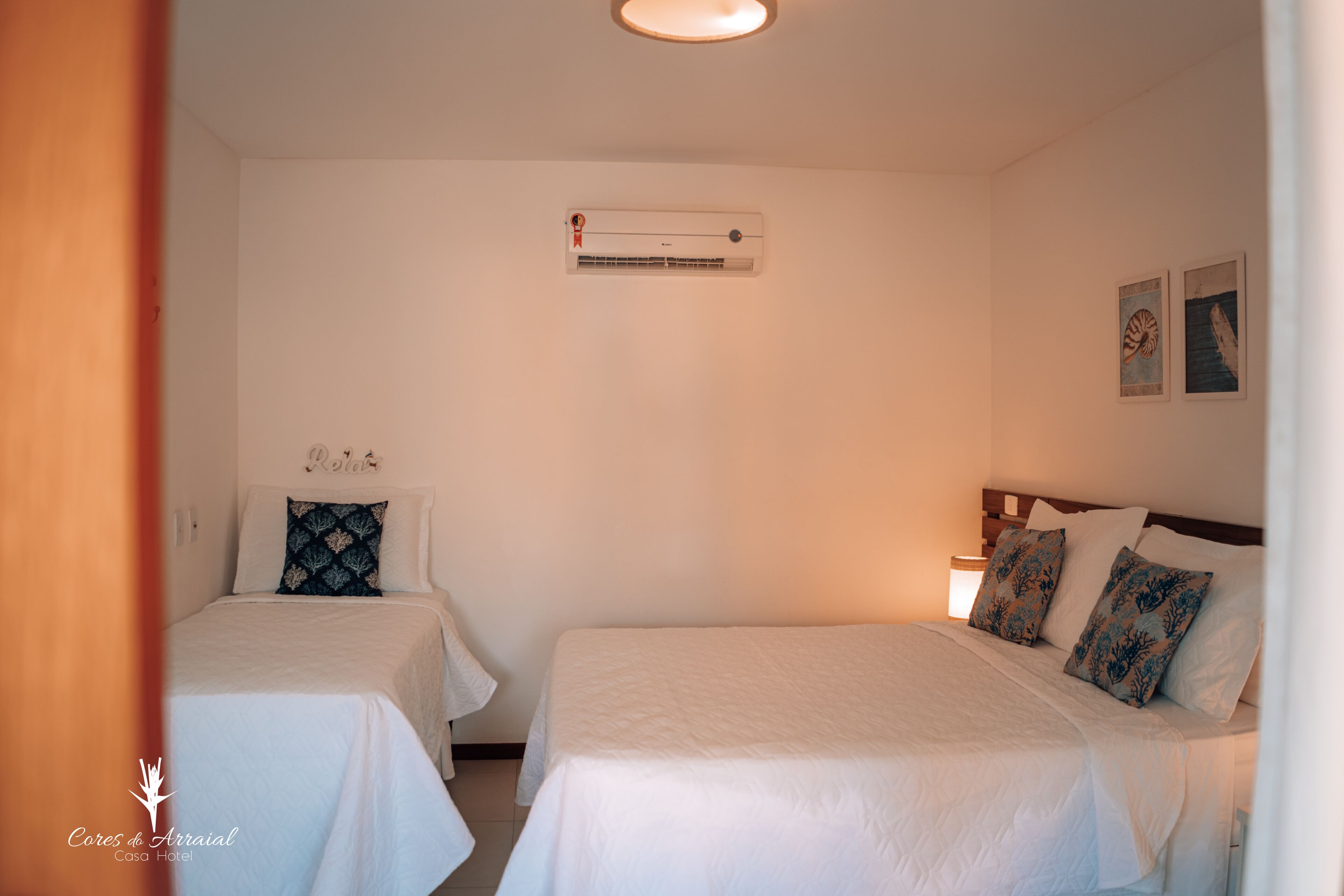 Habitación de hotel con dos camas, un ventilador de techo, un equipo de aire acondicionado y dos cuadros colgados en la pared.