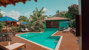 A pool area with lounge chairs and umbrellas, surrounded by tropical plants and a building with a tiled roof.