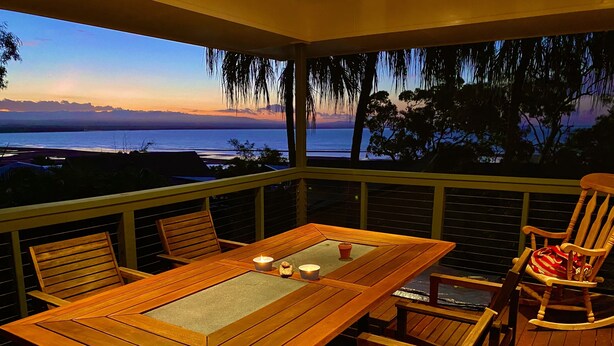 A wooden deck with a table and chairs, overlooking a body of water at sunset.