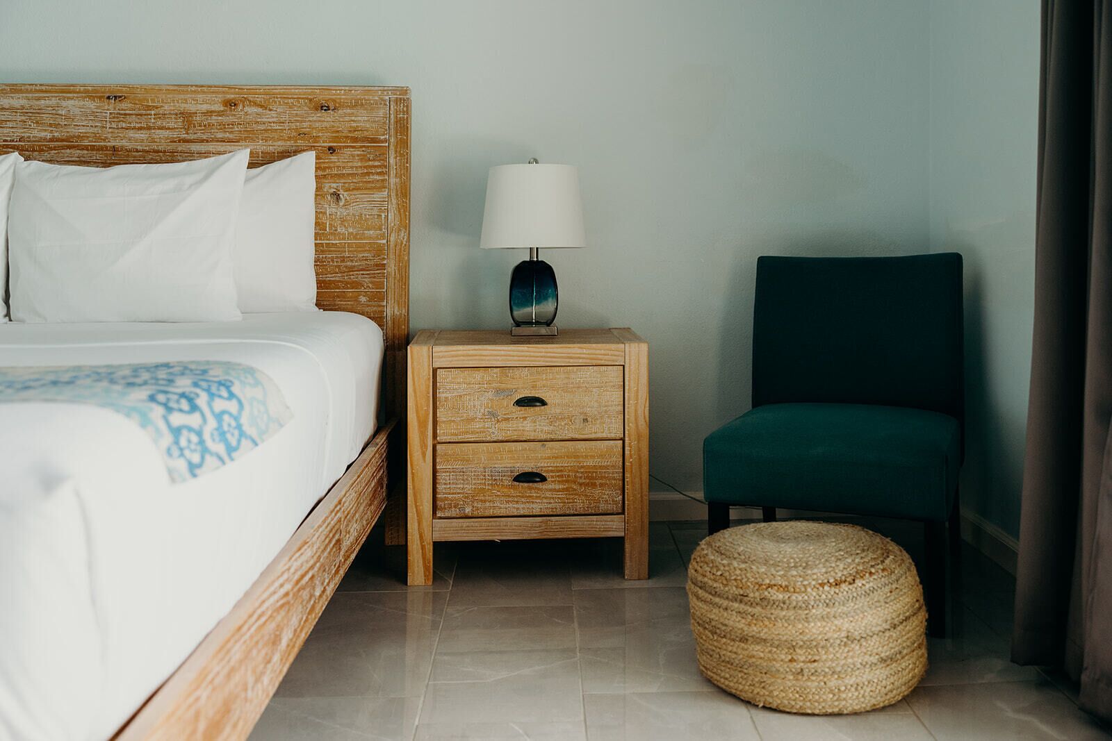 Une chambre d’hôtel avec un lit doté d’une tête de lit en bois, une table de chevet avec une lampe, un fauteuil vert et un pouf en osier.