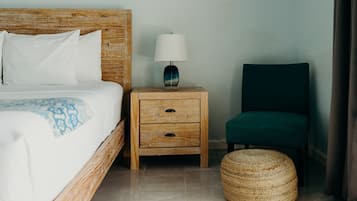 Une chambre d’hôtel avec un lit doté d’une tête de lit en bois, une table de chevet avec une lampe, un fauteuil vert et un pouf en osier.